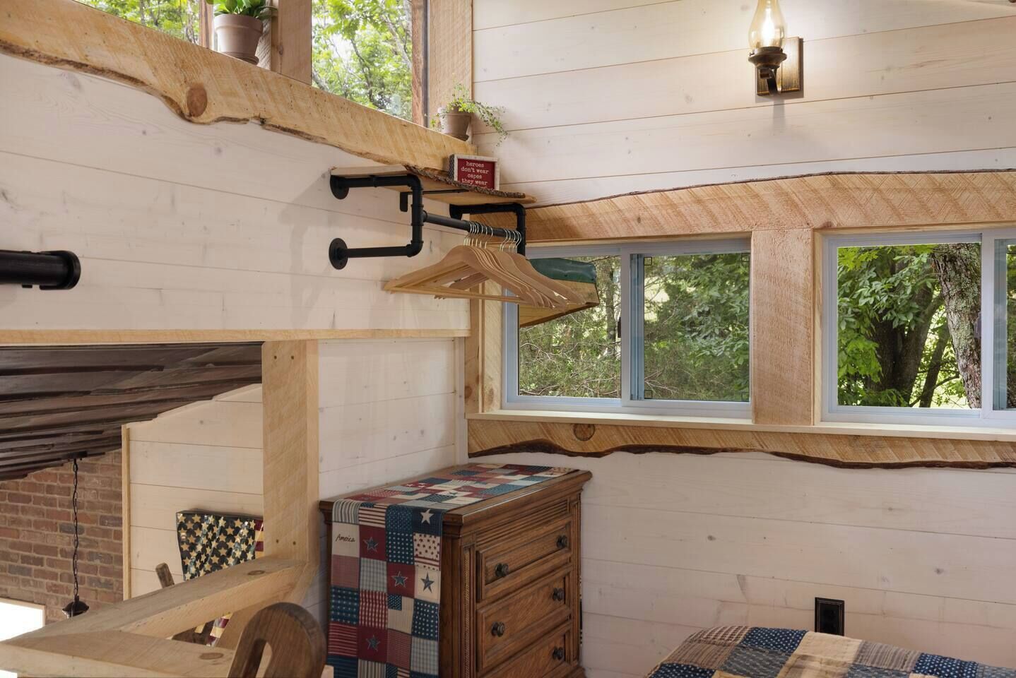 A rustic room with a wooden bench, a quilted chest of drawers, a window with a view of trees, and a sign on the wall.