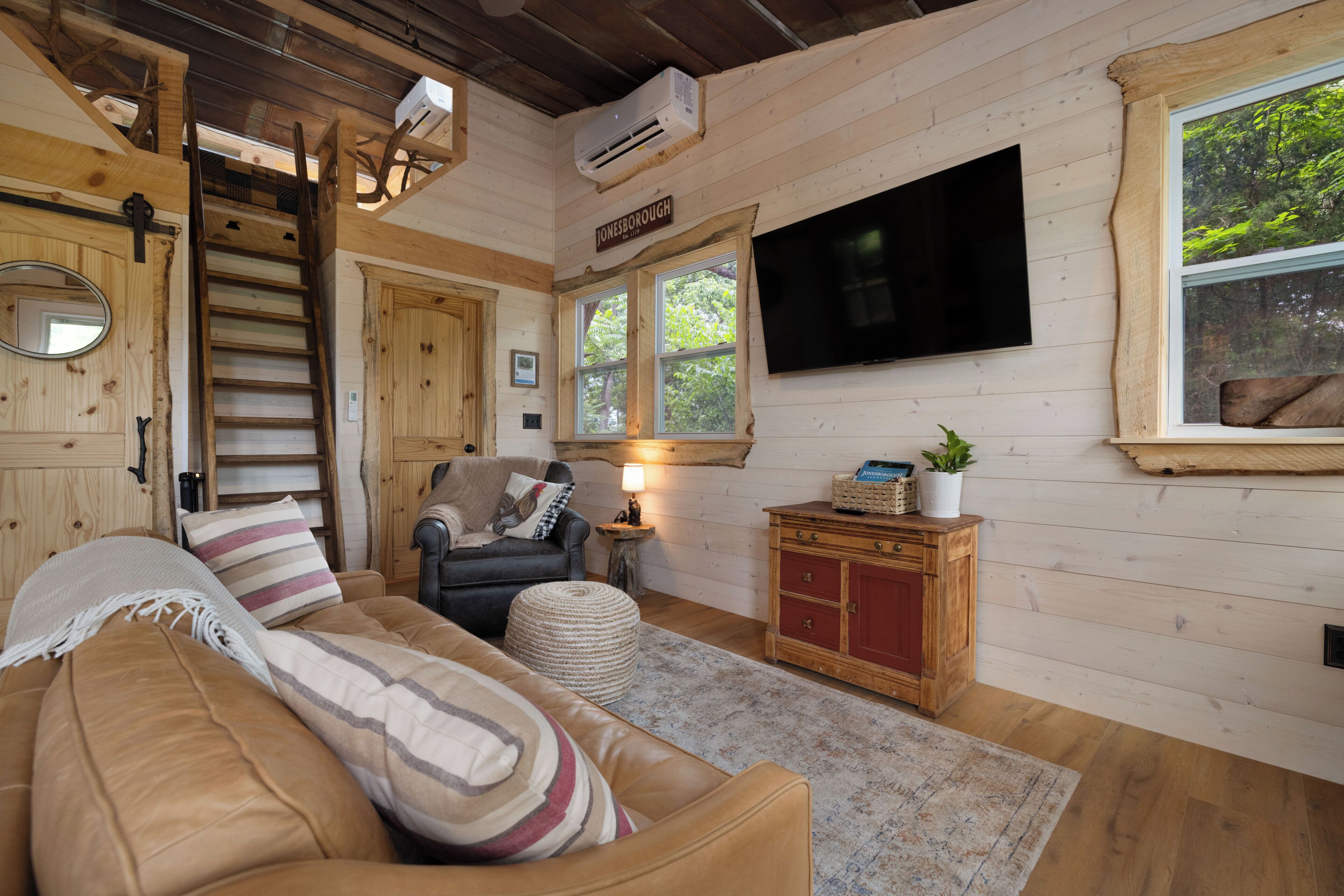 Een blokhut interieur met houten inrichting, een bank, een stoel, een televisie, een ladder en een raam met uitzicht op bomen.