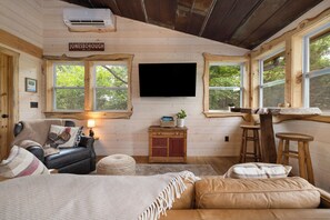 A cozy cabin interior with a leather sofa, a television mounted on the wall, wooden furniture, and large windows.
