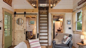 Interior acolhedor de uma cabana com porta de madeira, escada, sofá de couro, poltrona e uma pequena mesa com luminária.