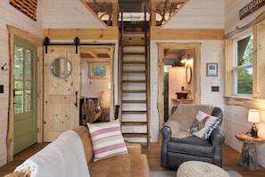 A cozy cabin interior with a wooden door, a ladder, a leather sofa, a chair, and a small table with a lamp.