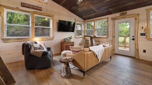 Interior de uma cabana de tronco com sofá, uma poltrona, uma mesinha, uma televisão e uma porta que dá para o exterior.