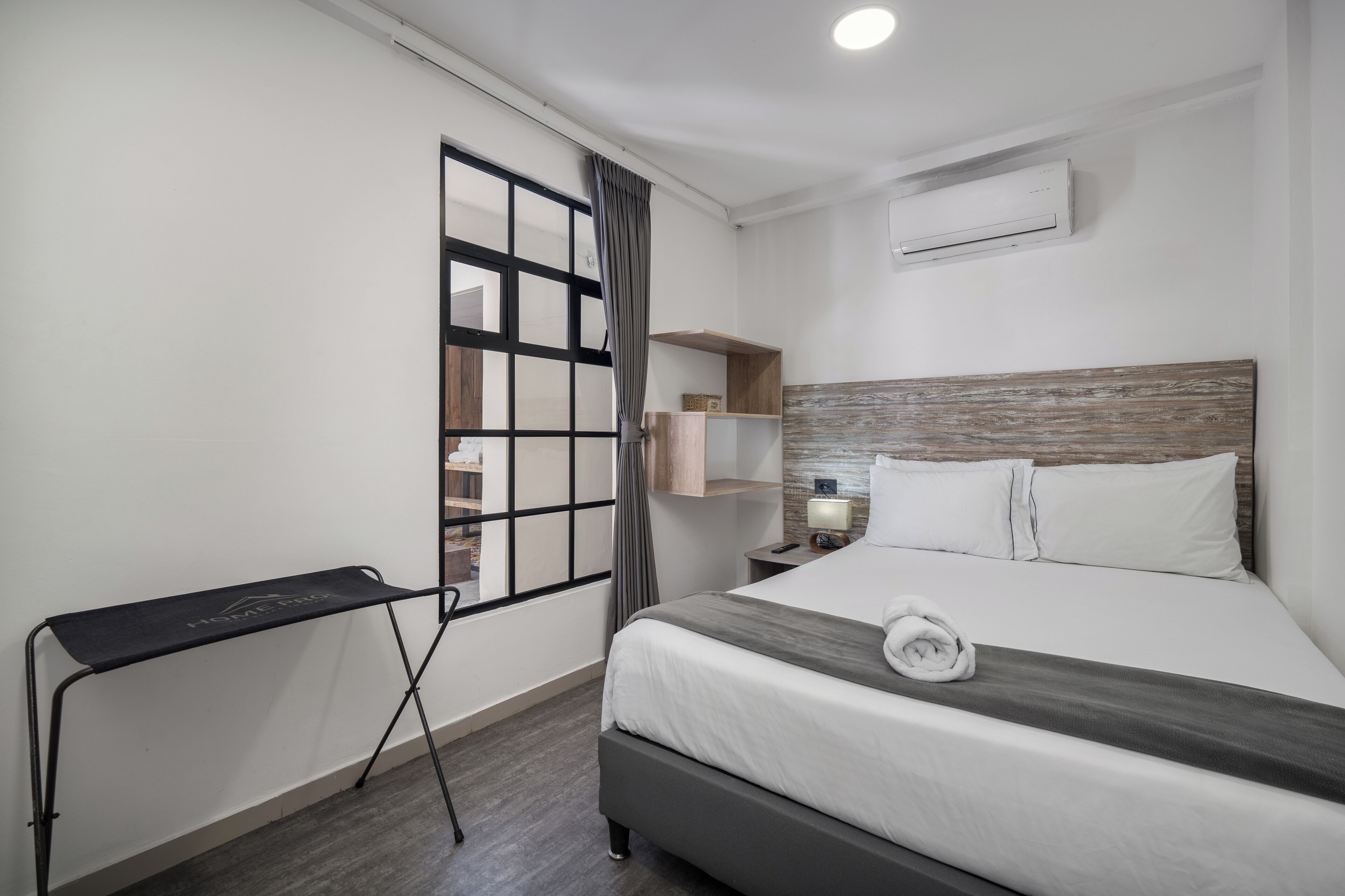 A hotel room with a bed, a desk, a window with curtains, a wall-mounted air conditioner, and a nightstand with a lamp.