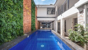 Una piscina con suelo de baldosas azules, rodeada por un muro de ladrillo con follaje y un edificio blanco con balcón.