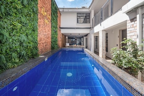 A swimming pool with a tiled blue floor, surrounded by a brick wall with greenery and a white building with a balcony. - Encanto House in Provenza by Nowa (Medellín)