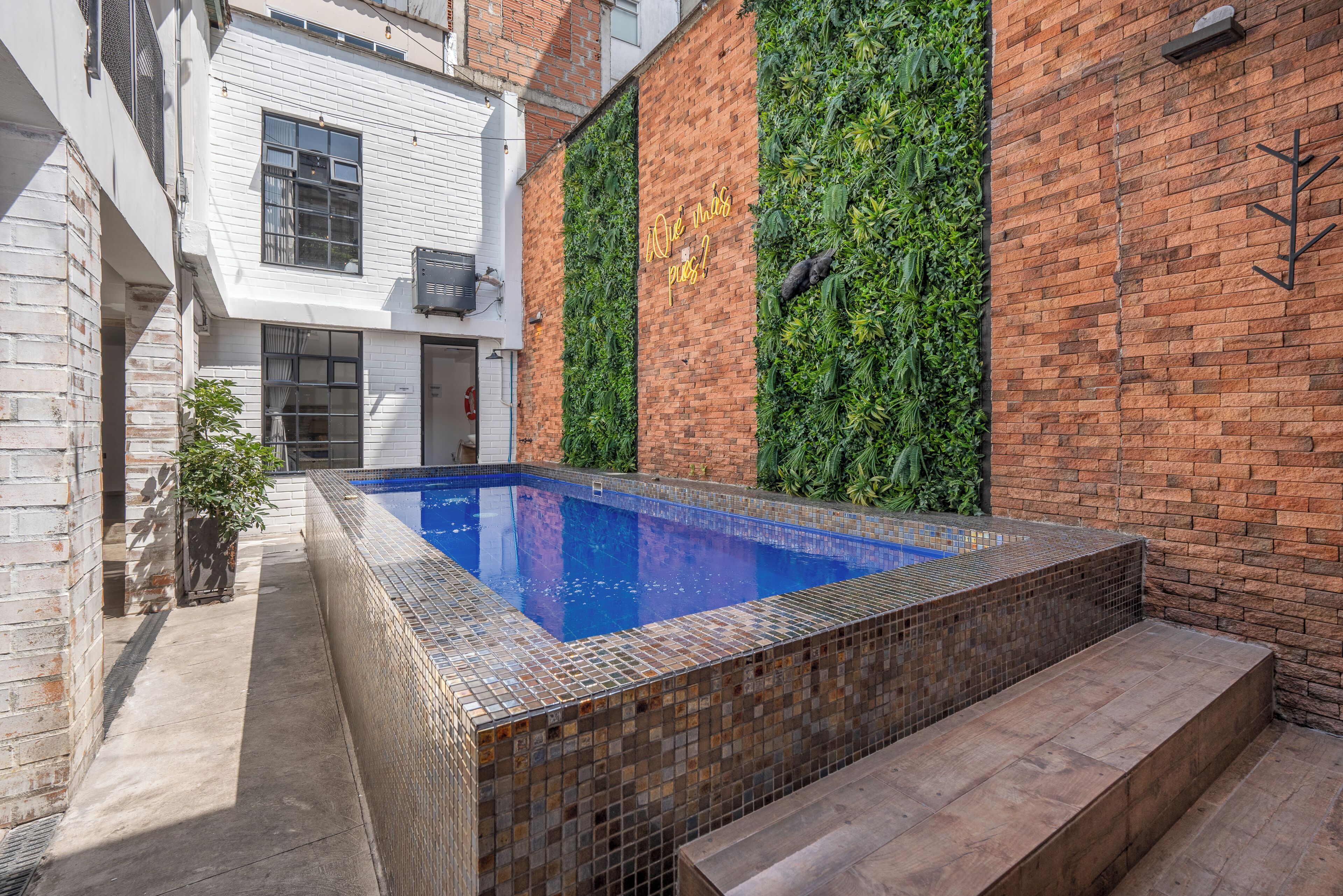 Une piscine dont le bord est recouvert de carreaux, entourée d’un mur de briques agrémenté de verdure et d’un bâtiment blanc doté de fenêtres.