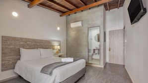 Un dormitorio moderno con cabecera de madera, cama con ropa blanca, unidad de aire acondicionado, mesita de noche con lámpara y televisor montado en la pared.