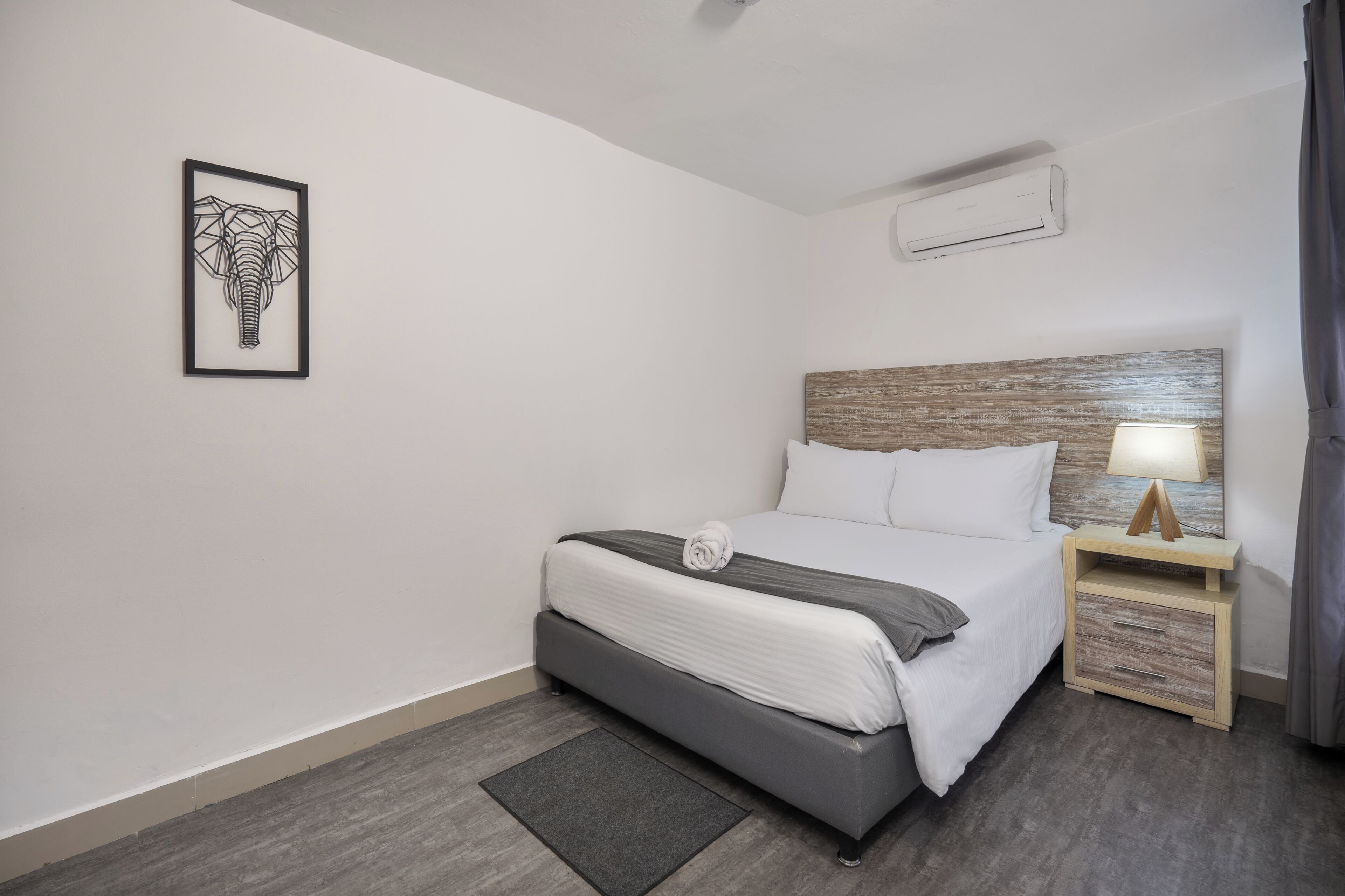 Habitación de hotel ordenada con cama, mesita de noche, lámpara, equipo de aire acondicionado y un cuadro enmarcado en la pared.