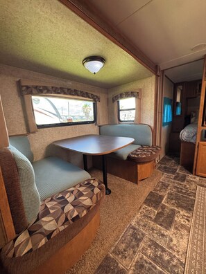 Ein kompaktes Wohnmobil-Interieur mit Essbereich, zwei gepolsterten Sitzgruppen, einem kleinen Tisch und einem Fenster mit Aussicht.