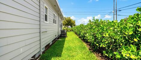 Une maison blanche avec un petit jardin devant, une pelouse verte et une rangée d’arbustes.