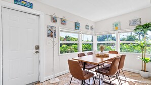 A dining area with a table and chairs, large windows with a view of greenery, and a white door. - NEW: Cozy Home w/ Hot tub & Beach Essentials! (Lake Worth Beach)