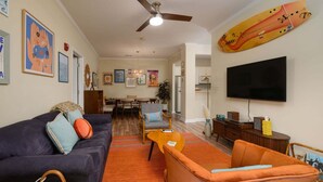 Ein Wohnzimmer mit Sofa, Sessel, Fernseher, Deckenventilator und Bildern an den Wänden.
