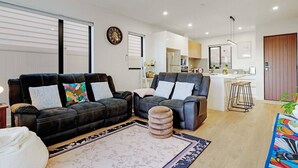 A modern living room with two sofas, a coffee table, and a kitchen in the background. - Spacious 4BR Family Haven with Air-con & Parking (Auckland South)
