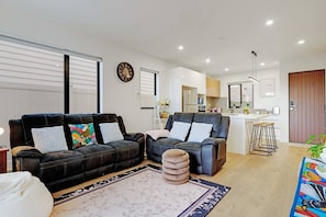 A modern living room with two sofas, a coffee table, and a kitchen in the background. - Spacious 4BR Family Haven with Air-con & Parking (Auckland South)