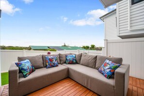 A beige sectional sofa with colorful pillows on a wooden deck, overlooking a green-roofed building.