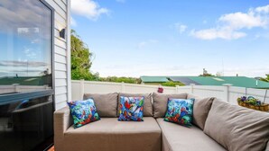 A sectional sofa with colorful pillows, a wooden deck, and a view of a green-roofed building in the background. - Spacious 4BR Family Haven with Air-con & Parking (Auckland South)