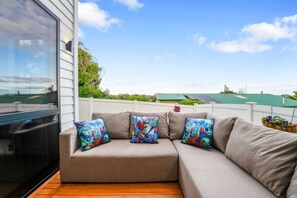 A sectional sofa with colorful pillows, a wooden deck, and a view of a green-roofed building in the background.