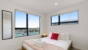 A bedroom with a bed, a nightstand, two windows, and a view of the cityscape. - Spacious 4BR Family Haven with Air-con & Parking (Auckland South)