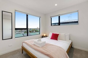 A bedroom with a bed, a nightstand, two windows, and a view of the cityscape.