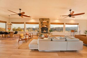A spacious living room with a stone fireplace, large windows, and ceiling fans.