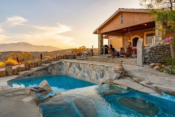 A pool with stone edges, a covered patio, and a view of mountains.