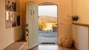 A hallway with a door leading to a desert landscape, a bench, and a plant on the wall. - Casa Sole | Pool | Spa | Near Palm Springs (Morongo Valley)