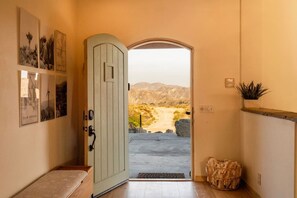 A hallway with a door leading to a desert landscape, a bench, and a plant on the wall.