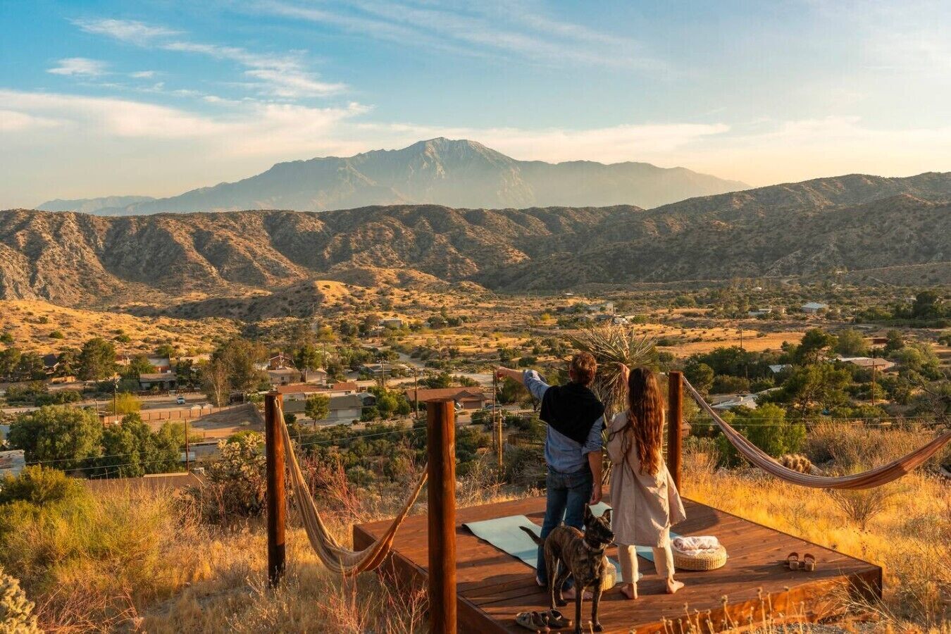 A couple standing on a wooden deck overlooking a mountainous landscape with a hammock and yoga mat.