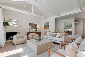 A spacious living room with a fireplace, a sofa, a coffee table, and a dining area visible through an open door. - NEW Montecito Hedgerow Jewel Box, 4BD/3BA, with Pool and Jacuzzi! (Santa Barbara)