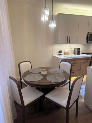 A dining area with a round table, four chairs, and a kitchen in the background.