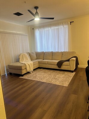 A living room with a sectional sofa, a ceiling fan, a window with sheer curtains, and a wooden floor.