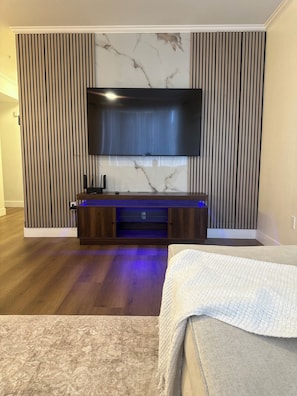 A modern living room with a flat-screen TV mounted on a wall with a marble-like design, a wooden entertainment unit with blue lighting, and a large bed with a white blanket.