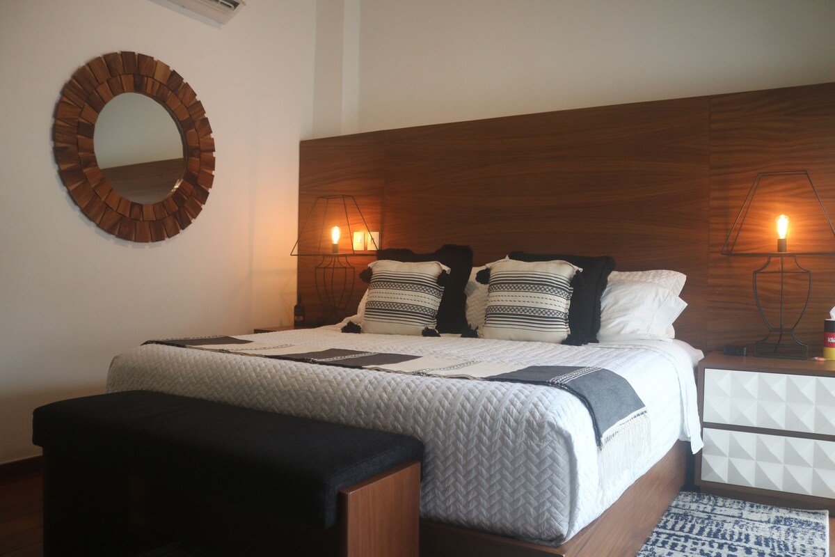 A bedroom with a wooden headboard, a round mirror, a bed with a quilted cover, two bedside lamps, and a bedside table with a lamp.