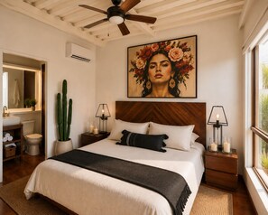 Een slaapkamer met een bed, een plafondventilator, een nachtkastje met een lamp, een pot met een cactus en een groot bloemen schilderij aan de muur.