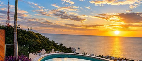 Ein Schwimmbecken mit Blick auf den Sonnenuntergang über dem Meer, umgeben von Grünflächen und einer Holzterrasse.