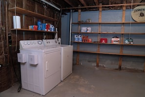 A laundry room with a washer and dryer, shelves with cleaning supplies, and a wall-mounted sign. - Charming Cape with AC near downtown Falmouth (Falmouth)