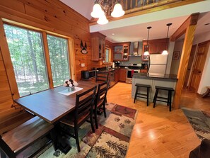 A wooden cabin interior with a dining table, chairs, a kitchen area, and a large window overlooking a forest. - The Elkstone (Long Lake)