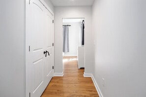 Ein schmaler Korridor mit weißen Wänden und Holzboden, der zu einem Zimmer mit einem Bett und einem fensterförmigen Durchbruch mit Vorhängen führt.