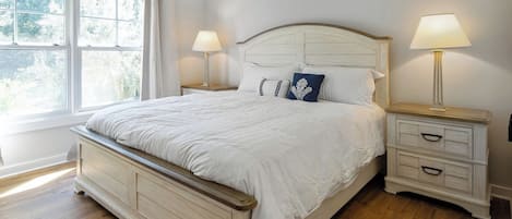 Un dormitorio con una cama grande, dos mesitas de noche con lámparas, un ventanal con cortinas y suelo de madera.