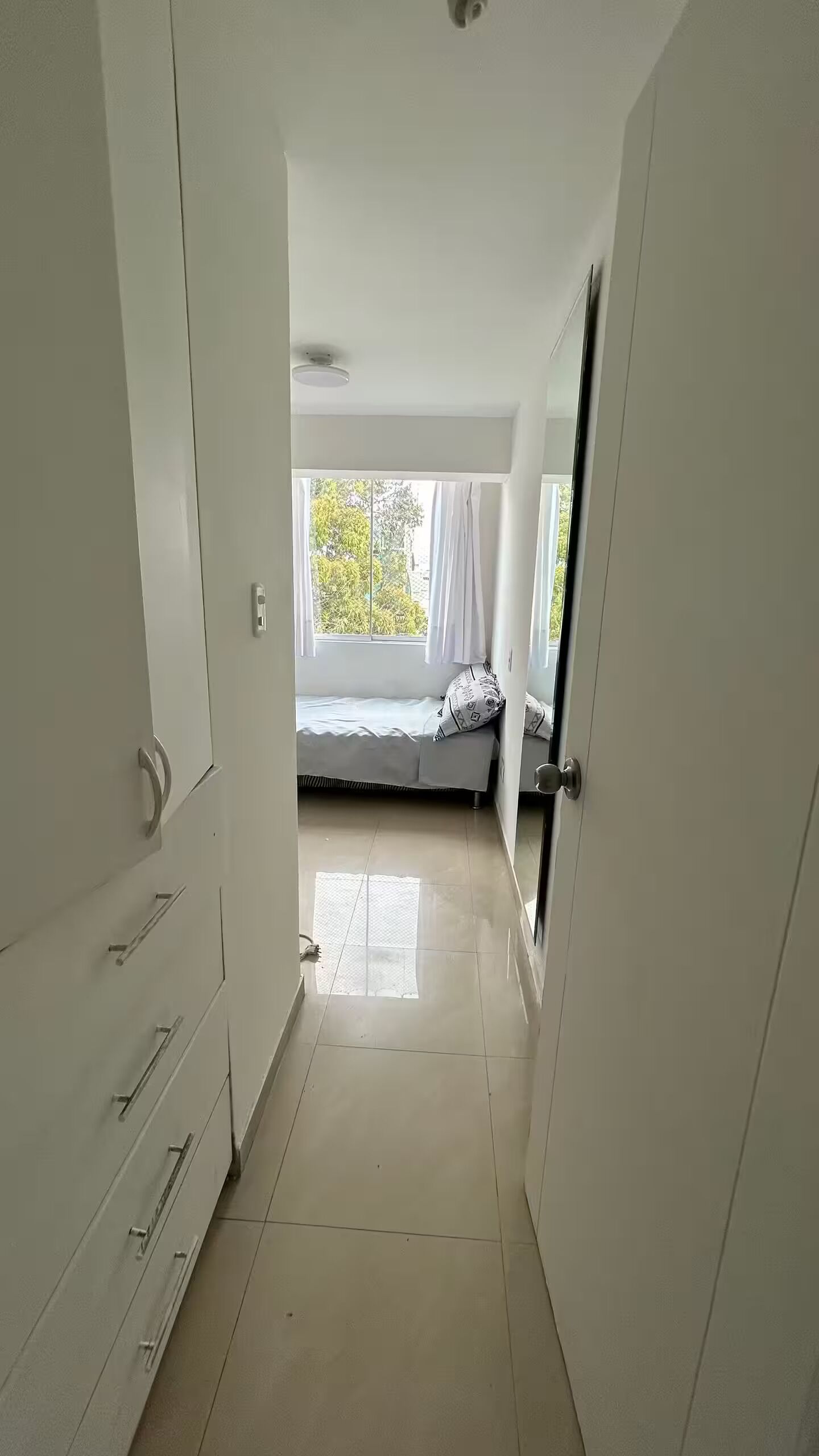 A corridor leading to a bedroom with a bed, a window, and a door.