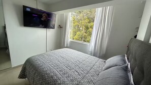 Una camera da letto con un letto, una TV a parete, una finestra con tende e vista sugli alberi.