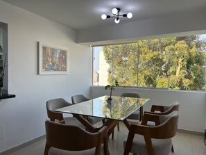 Zona pranzo con tavolo in vetro, sedie in legno, lampadario a soffitto e finestra vista alberi.