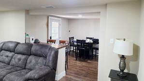 Un salon avec un canapé en cuir, un coin repas avec une table et des chaises, une lampe et une porte donnant sur une autre pièce.