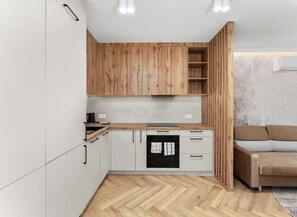 A compact kitchen with wooden cabinets, a built-in oven, and a grey sofa in the adjacent living area. - City Łobzów Harmony Apartment (Kraków)