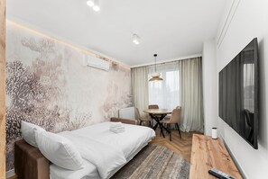 Una habitación de hotel moderna con una cama grande, una mesa de comedor, sillas, un televisor y un aire acondicionado.