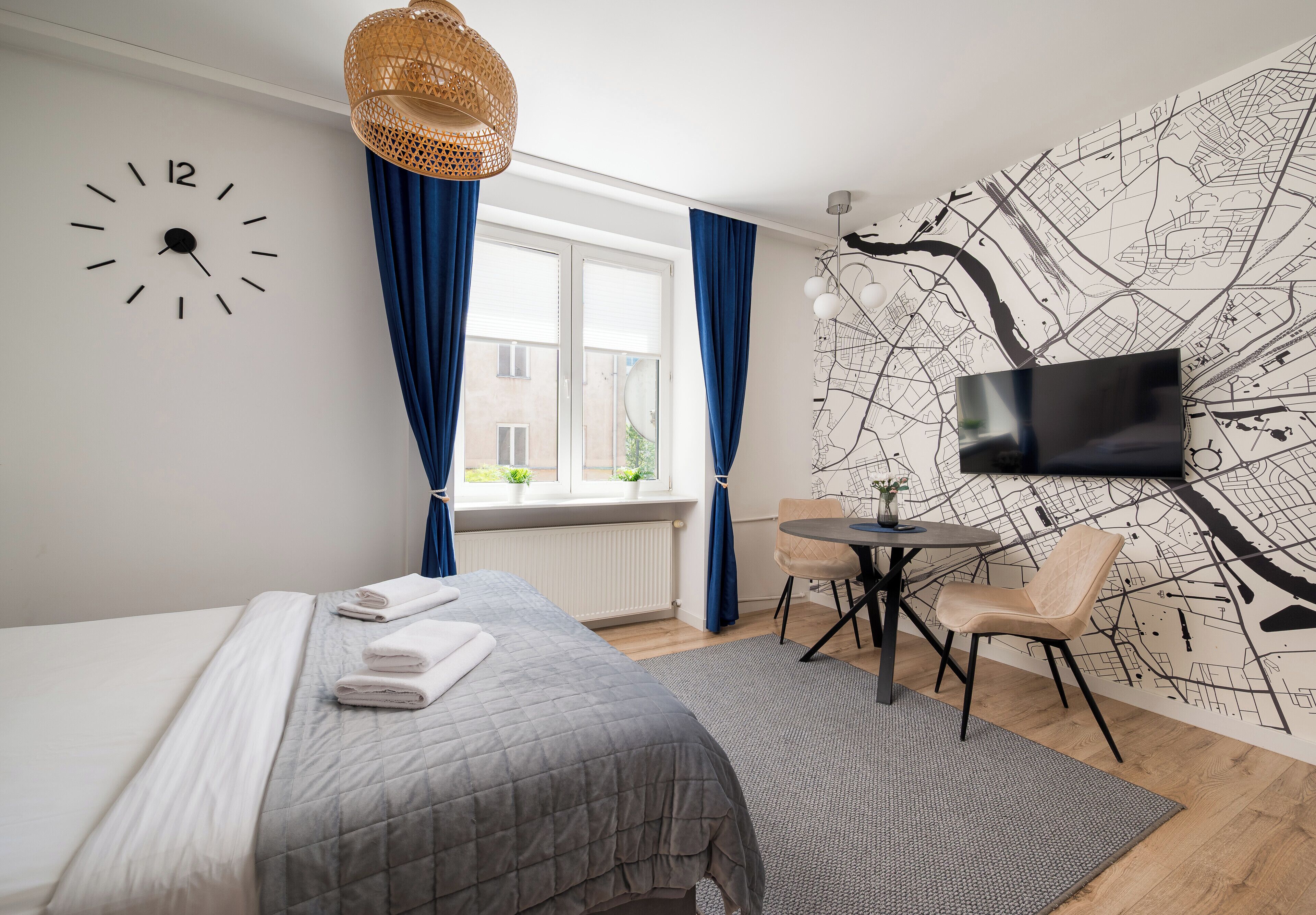 Ein modernes Hotelzimmer mit Bett, Wanduhr, Fernseher, Esstisch mit Stühlen und einer Wandmalerei mit Stadtkarte.