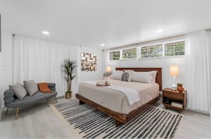Ein Schlafzimmer mit einem Bett, Nachttischen, einem Sofa, einer Zimmerpflanze und einem Fenster mit Vorhängen.