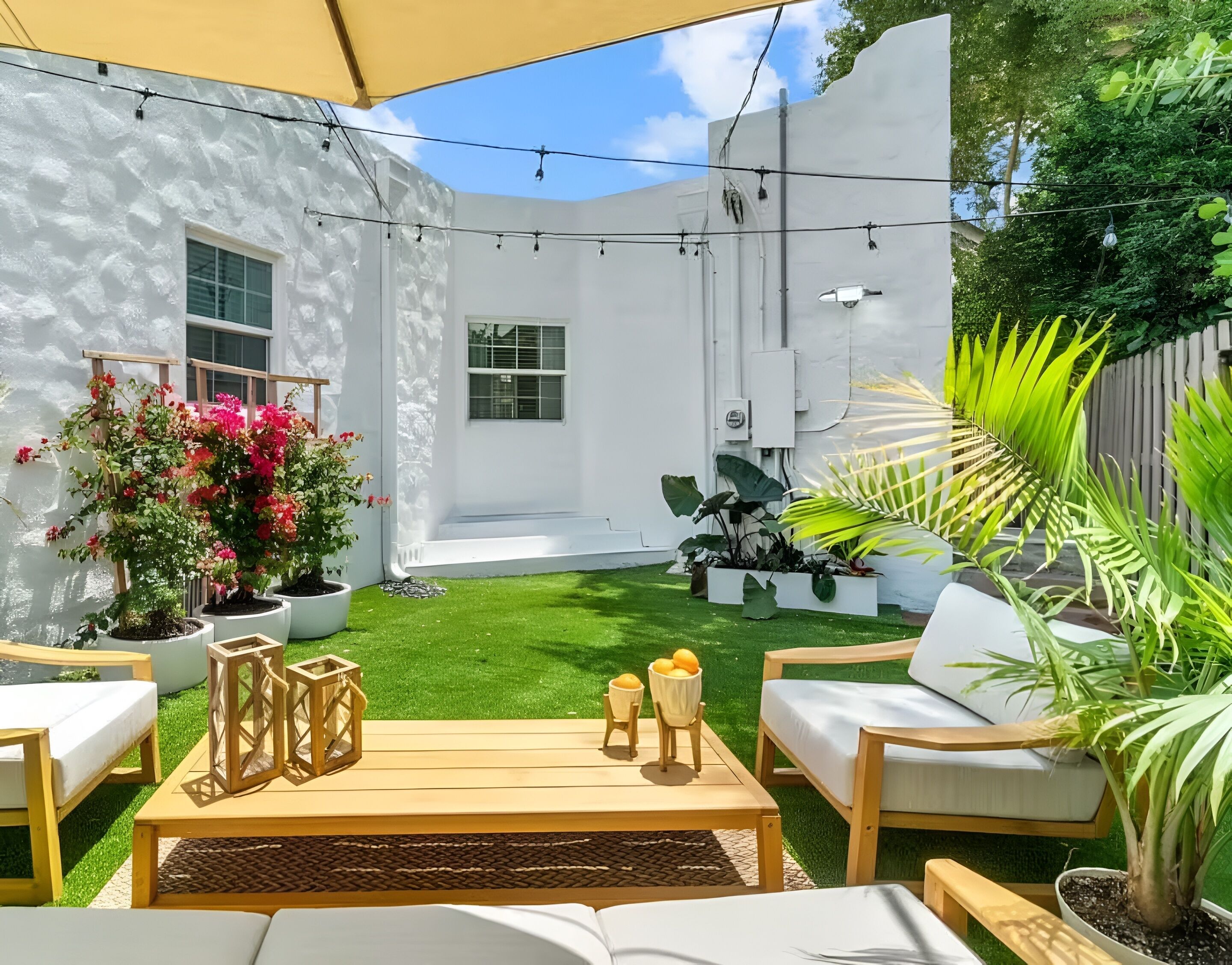 A patio area with a white wall, a wooden table, two chairs, a small table with candles, and potted plants.
