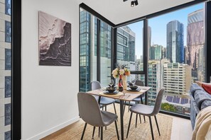 A dining area with a table set for two, a city view through large windows, and a modern interior design. - CBD-Edge 2BR | Pool, Gym, Sauna & City Views (West Melbourne)