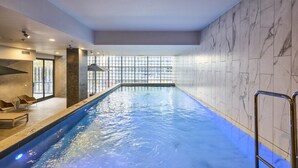 An indoor swimming pool with a clear view of the outside through large windows, a poolside seating area, and a modern interior design. - CBD-Edge 2BR | Pool, Gym, Sauna & City Views (West Melbourne)